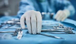 A gloved hand reaches for a surgical instrument on a table covered with various metal tools. The person is wearing blue surgical attire, following ANSI/AAMI ST108:2023 Standards, and the background is slightly out of focus, suggesting a medical or surgical setting.